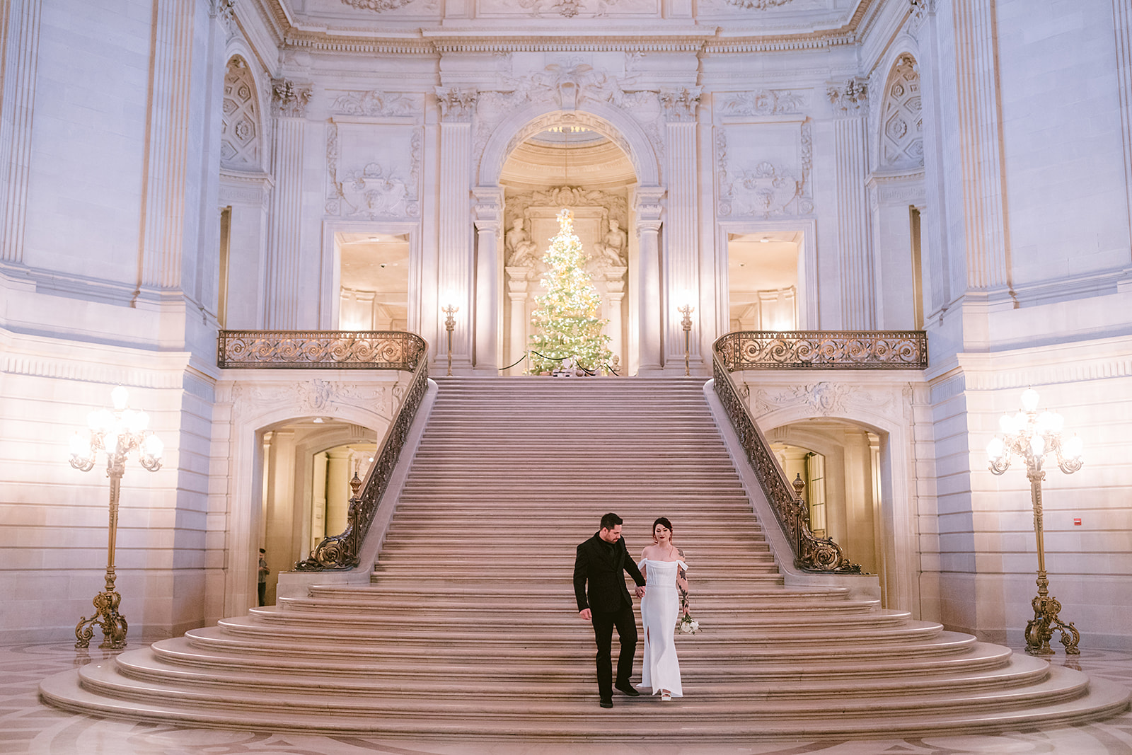 Bride and groom walking down the grand staircase at San Francisco City Hall with a glowing Christmas tree behind them – elegant and romantic San Francisco elopement photography